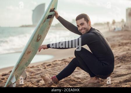 Vista laterale dell'uomo in muta nera seduta sulla spiaggia di sabbia con tavola da surf che si affaccia in contemplazione, Spagna Foto Stock