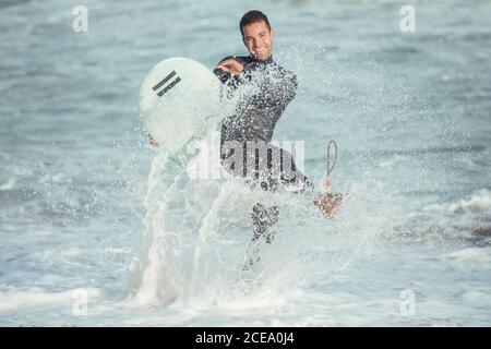 Elettrizzato uomo sportivo con tavola da surf che spruzzi d'acqua e ridendo alla macchina fotografica alla luce del sole, Barcellona Foto Stock
