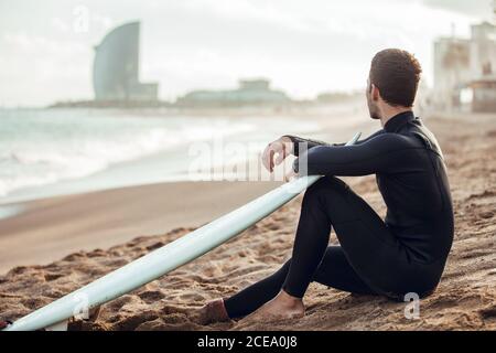 Vista laterale dell'uomo in muta nera seduta sulla spiaggia di sabbia con tavola da surf che si affaccia in contemplazione, Spagna Foto Stock