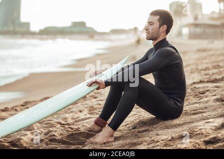 Vista laterale dell'uomo in muta nera seduta sulla spiaggia di sabbia con tavola da surf che si affaccia in contemplazione, Spagna Foto Stock