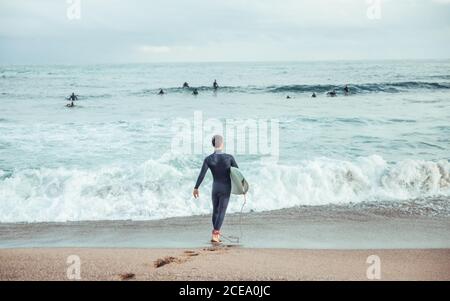 Vista posteriore di uomo in muta correndo su onde ruvide dell'oceano che porta tavola da surf, Spagna Foto Stock