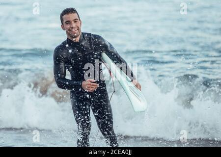 Elettrizzato uomo sportivo con tavola da surf che spruzzi d'acqua e ridendo alla macchina fotografica alla luce del sole, Barcellona Foto Stock