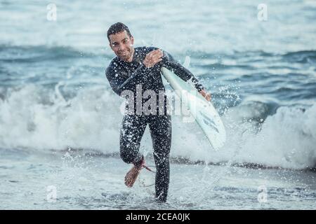 Elettrizzato uomo sportivo con tavola da surf che spruzzi d'acqua e ridendo alla macchina fotografica alla luce del sole, Barcellona Foto Stock
