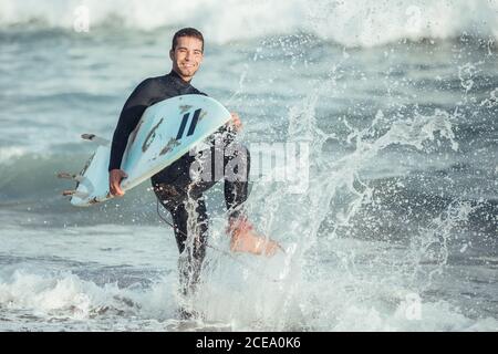Elettrizzato uomo sportivo con tavola da surf che spruzzi d'acqua e ridendo alla macchina fotografica alla luce del sole, Barcellona Foto Stock