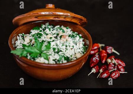 Un mucchio di peperoncini rossi caldi che si trova vicino alla ciotola in ceramica con gustosa cagliata con erbe su un tavolo scuro Foto Stock