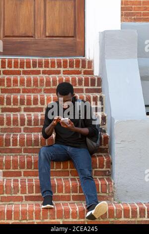 Annapolis, MD 08/21/2020: Un giovane afroamericano che indossa una t-shirt nera, jeans, sneakers e uno zaino è seduto su scale di mattoni fuori una h. Foto Stock
