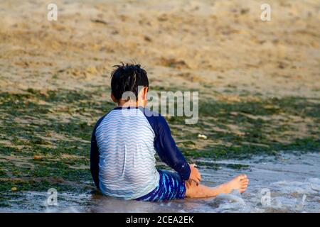 Un ragazzo caucasico giovane che indossa costume da bagno a manica lunga di protezione dell'eruzione è seduto sulla sabbia vicino al mare. Sta giocando da solo con la sabbia e l'acqua. L'immagine era ta Foto Stock