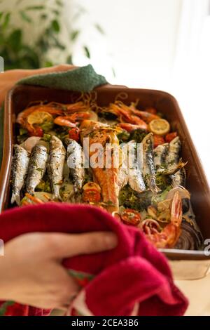 Mani di anonima Donna prendere pentola in ceramica con gustoso piatto di pesce dal forno caldo a casa Foto Stock