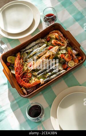 Padella con piatti di pesce e bicchieri di vino rosso in piedi su una tovaglia a scacchi Foto Stock