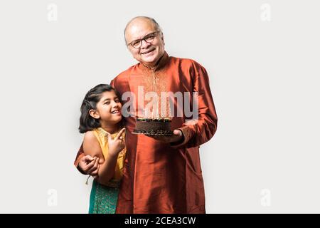 L'anziano o l'anziano indiano con la nipote che festeggia il compleanno soffiando le candele sulla torta mentre indossa l'usura etnica, in piedi isolato contro il bianco Foto Stock