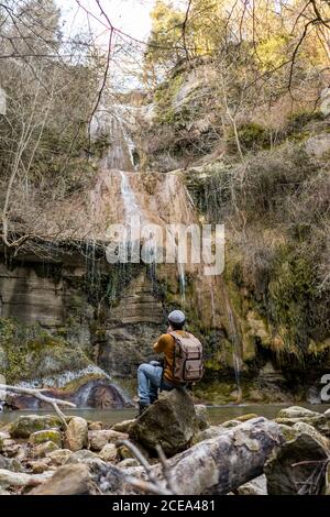 Vista posteriore del viaggiatore maschio con seduta sulle pietre e scattare foto con la fotocamera del fantastico cascata nella foresta in Spagna Foto Stock