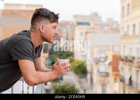 Vista laterale di un uomo informale che tiene una tazza di caffè e. appoggiandosi su una recinzione di balcone che guarda lontano Foto Stock