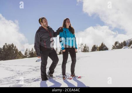 Allegro uomo e donna in abiti caldi e racchette da neve sorridendo e guardando lontano mentre si è in piedi sulla neve nella meravigliosa natura invernale Foto Stock