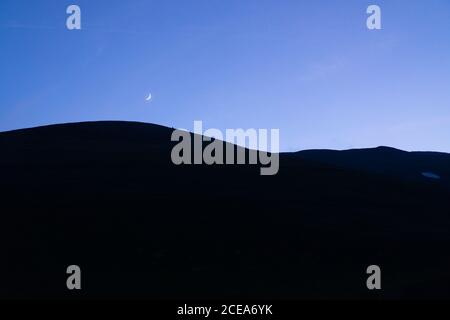 Paesaggio minimalista di silhouette nere di colline di montagna contro il crepuscolo cielo blu con luna crescente Foto Stock