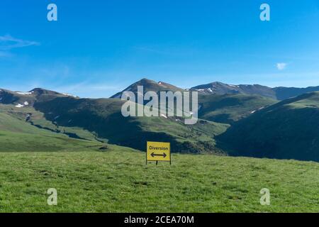 Paesaggio verde di montagne e segno giallo che mostra diversione con frecce in direzioni diverse sotto il cielo blu Foto Stock