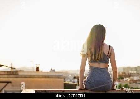 Vista posteriore della giovane brunetta FIT Donna in abito estivo alla moda seduta rilassata sul parapetto della terrazza sul tetto del vecchio edificio con la città sfocata su sfondo retroilluminato Foto Stock