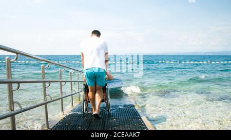 Uomo disabile in spiaggia che nuota su una sedia a rotelle con assistenza su una rampa accessibile. Foto Stock