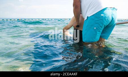 Uomo disabile in spiaggia che nuota su una sedia a rotelle con assistenza su una rampa accessibile. Foto Stock