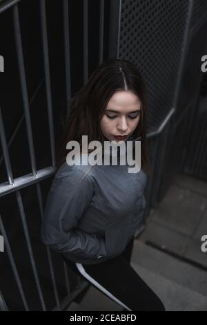Dall'alto appassionata giovane donna in abbigliamento sportivo con trucco e. mani in tasche in piedi sulle scale vicino alla griglia di acciaio Foto Stock