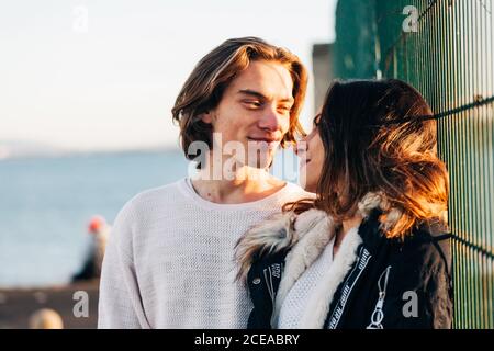 Uomo compiaciuto e donna felice attraente in cappotto che si guardano vicino a recinzione di metallo Foto Stock