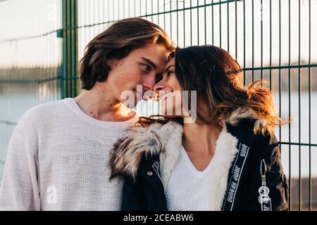 Uomo compiaciuto e donna felice attraente in cappotto che si guardano vicino a recinzione di metallo Foto Stock