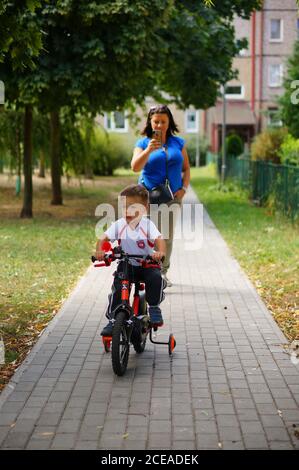 POZNAN, POLONIA - 30 agosto 2020: Piccolo bambino che guida una piccola bicicletta con ruote di supporto su un sentiero. Donna che segue il ragazzo. Foto Stock