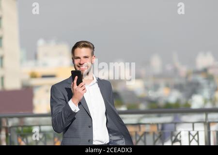 Imprenditore sorridenti utilizza lo smartphone per le video chiamate o messaggi di testo, skyline background. Uomo in tuta imprenditore assume i vantaggi delle moderne tecnologie mobili per il business. Business il concetto di chiamata. Foto Stock