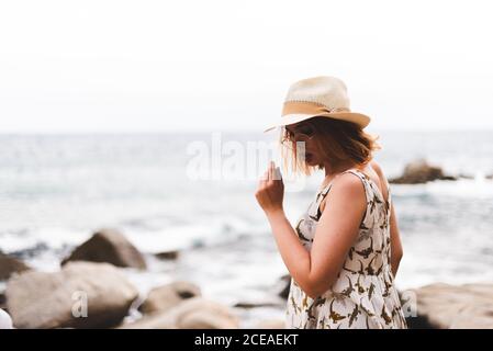 Vista posteriore della donna tatuata in cappello in piedi sulle pietre con le mani a parte e guardando l'oceano Foto Stock