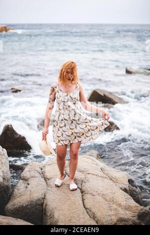 Vista posteriore della donna tatuata in cappello in piedi sulle pietre con le mani a parte e guardando l'oceano Foto Stock