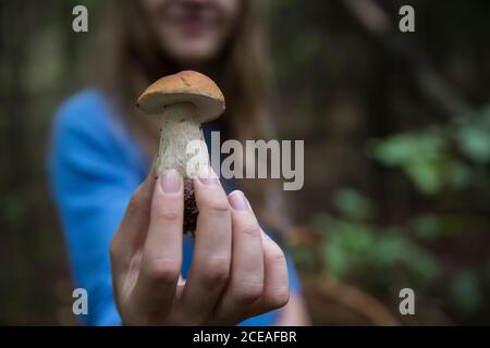 Bella giovane donna con cestino dimostrando piccolo fungo alla macchina fotografica mentre in piedi nella foresta verde Foto Stock