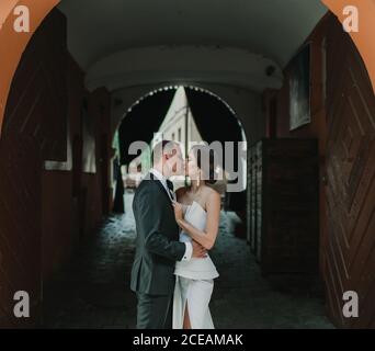 Vista laterale di giovane sposa e ginestra abbracciando e baciando l'uno con l'altro mentre si trova nell'arco di un vecchio edificio via della città Foto Stock