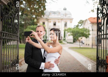 Bella giovane sposa e bello sposo sorridente e utilizzando smartphone per prendere selfie vicino al cancello d'ingresso del giardino di lusso Foto Stock
