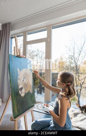 Vista laterale di pittura di disegno di ragazza piccola di talento seduto a. cavalletto contro la finestra a casa Foto Stock