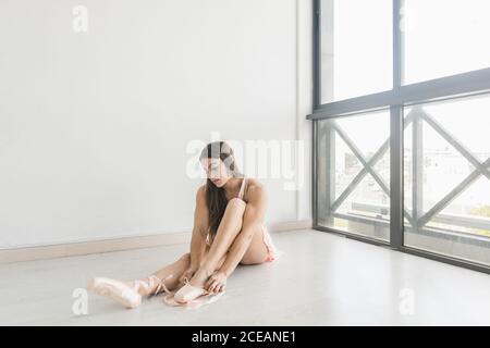 Giovane donna sottile e attraente che indossa le scarpe da ballo e si siede sul pavimento in una stanza luminosa vicino alla finestra Foto Stock
