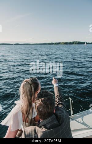 Vista posteriore dello sposo che punta via mentre si è in piedi con la sposa in barca sul lago Foto Stock