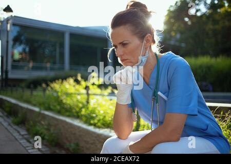 covid-19 pandemic. pensive medico moderno donna in scrub con stetoscopio e maschera medica seduta all'aperto vicino ospedale. Foto Stock