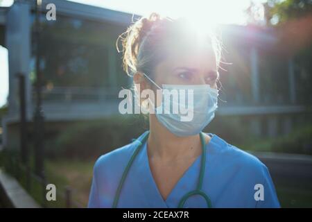 pandemia di coronavirus. triste donna medica moderna in scrub con stetoscopio e maschera medica all'esterno vicino alla clinica. Foto Stock