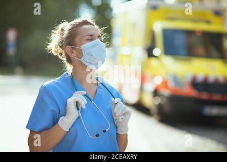 pandemia di coronavirus. donna moderna paramedica stanca in uniforme con stetoscopio e maschera medica che respira all'esterno vicino all'ambulanza. Foto Stock