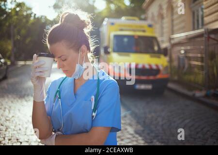 covid-19 pandemic. stanco medico donna moderna in uniforme con stetoscopio, maschera medica e tazza di caffè fuori vicino ambulanza. Foto Stock
