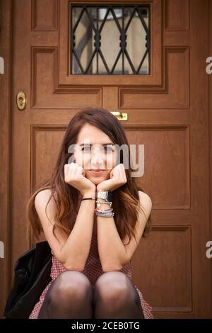 Giovane donna attraente in abito con braccialetti a mano e capelli scuri seduti sul portico vicino a porta di legno di casa e guardando la macchina fotografica Foto Stock