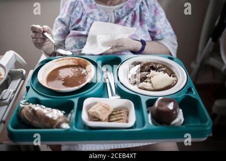 Donna matura irriconoscibile ricoverata in ospedale per la chirurgia del ginocchio, seduta sulla sedia, mangiare cibo dal vassoio Foto Stock