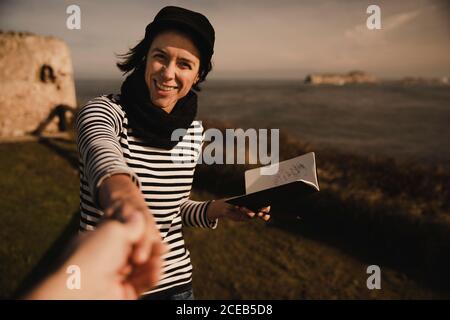 Elegante lady allegro nel cappuccio con notepad tenendo la mano del raccolto di persona sulla costa vicino al mare Foto Stock