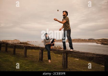 Giovane ragazzo in hat juggling balls vicino elegante signora nel cappuccio con tamburo etica seduto sul sedile vicino alla costa del mare e cielo molto nuvoloso Foto Stock