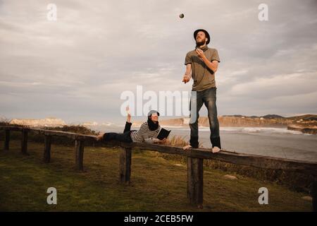 Giovane ragazzo in hat juggling balls vicino elegante signora nel cappuccio con tamburo etica seduto sul sedile vicino alla costa del mare e cielo molto nuvoloso Foto Stock