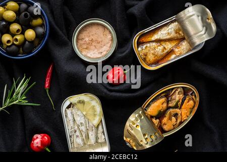 Ciotola con olive e vari peperoni caldi che si trovano vicino a scatola pesce su fondo di stoffa nera Foto Stock