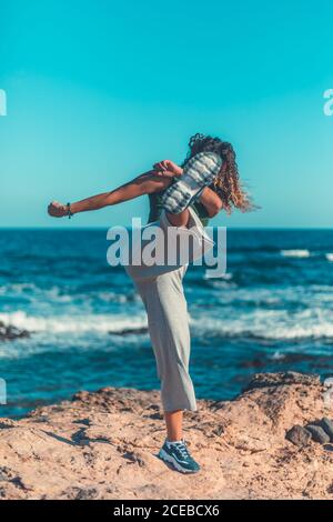 Irriconoscibile femmina in abito casual calci aria mentre in piedi sulla riva sassosa contro agitando il mare e cielo blu senza nuvole Foto Stock