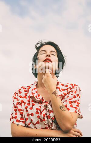 Dal basso vista di attraente brunetta Donna in camicia a motivi con tatuaggi sul braccio in piedi sullo sfondo di cielo nuvoloso con gli occhi chiusi Foto Stock