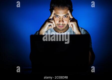 Giovane ragazzo sfregando le tempie con le dita e guardando la macchina fotografica seduto vicino al computer portatile su sfondo blu Foto Stock