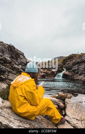 Vista posteriore di signora in cappello e impermeabile giallo seduta su pietre spaccate di fiume tra colline rocciose utilizzando il telefono cellulare a Isoba, Castiglia e Leon, Spagna Foto Stock