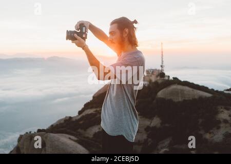 Vista laterale di un bel ragazzo bearded utilizzando una fotocamera professionale per scattare foto su una natura incredibile mentre si alza in montagna durante l'alba a Barcellona, Spagna Foto Stock